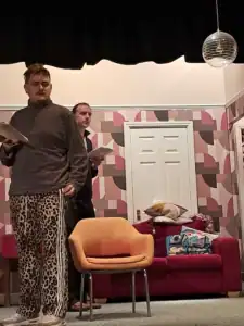 Two men reading scripts on stage in a colourful room setting.